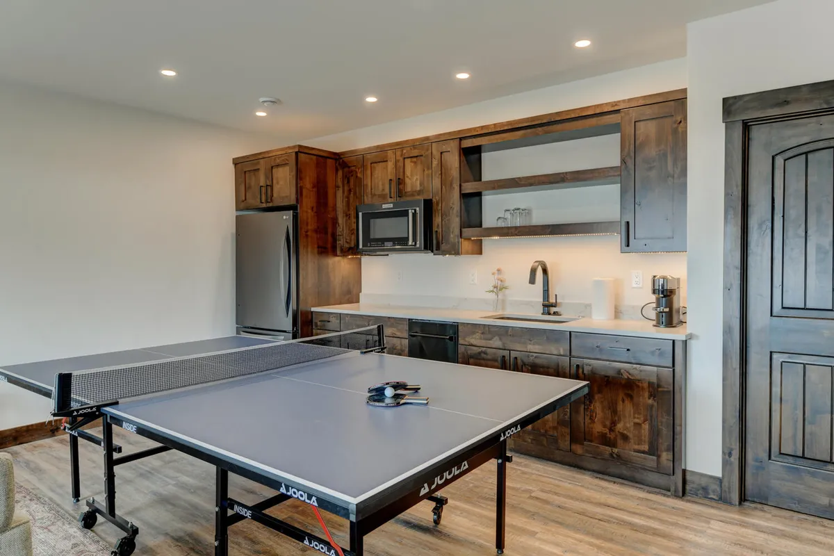 An inviting basement area with a ping pong table, dark wood cabinetry, and modern kitchen appliances. The scene sets the stage for both playful activity and culinary convenience, with ample space for group gatherings.