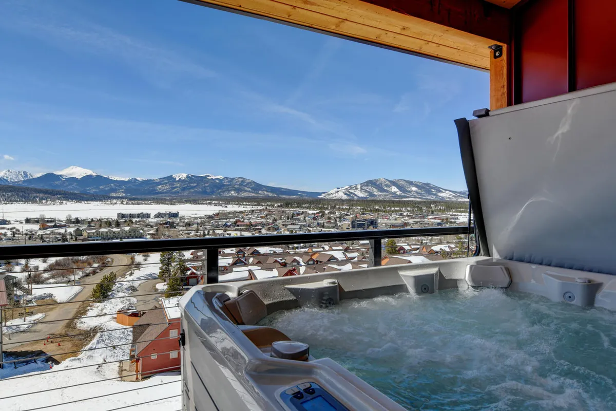 This image captures a tranquil hot tub on a private deck overlooking a snowy mountain landscape, with the Rockies stretching into the distance. The scene shows guests relaxing in the steaming water, surrounded by fresh alpine air and serene views, with hints of socializing and stargazing.