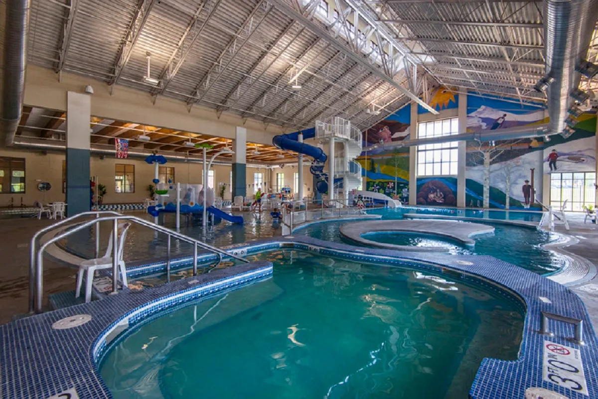 A colorful, energetic interior of a recreation center pool area, featuring water slides, a lazy river, children playing, and vibrant murals on the walls. The scene is full of movement and family fun.