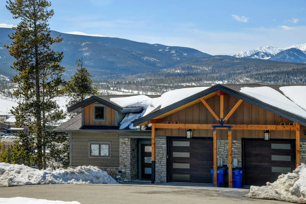 A striking exterior view of the villa features stone and wood architecture set against a majestic mountain backdrop. The front of the home is inviting with garage doors, a spacious driveway, and hints of landscaped outdoor space—offering the perfect launching point for outdoor adventures with pets.
