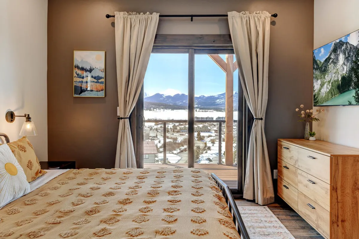 This serene bedroom features a bed with a patterned coverlet positioned beside a large window that frames spectacular mountain views. Decorative artwork and a wooden dresser enhance the inviting ambiance, while a potted plant adds a touch of calm. The space feels tranquil and perfectly suited for restful retreats.