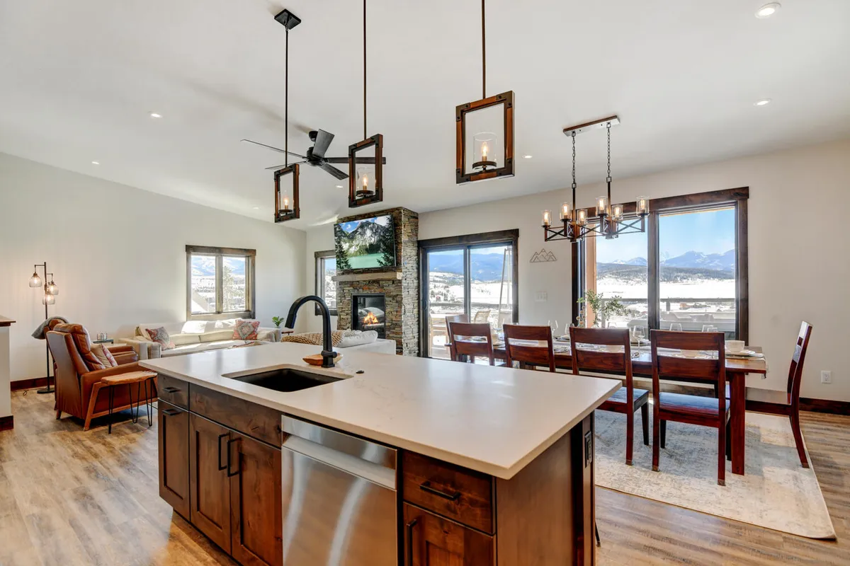 An open-concept interior blends the kitchen, dining area, and great room, featuring a large stone fireplace, expansive windows with mountain views, and comfortable seating. The image radiates warmth, offering a glimpse of multiple gathering spots within the heart of the home.