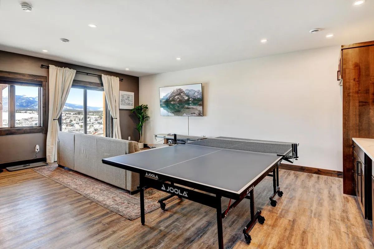 A lively, well-lit entertainment den featuring a ping pong table at the center, a cozy sofa for relaxation, a large window with scenic views, and playful mountain artwork. The space is set up for both active games and relaxed group gatherings.