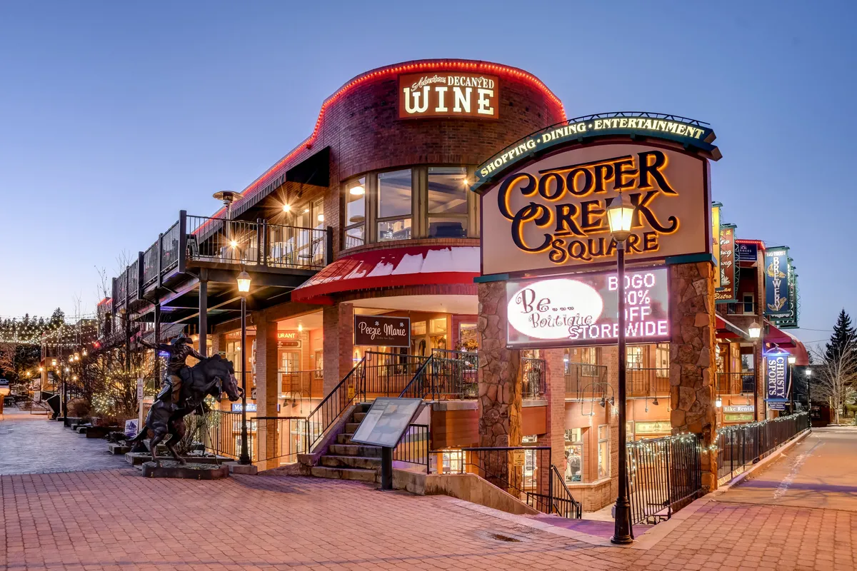 An exterior view of Cooper Creek Square, showcasing a lively public space with signage for shopping and wine, outdoor seating, and a horse statue. The festive, welcoming mood reflects a vibrant local hub for dining, shopping, and entertainment.