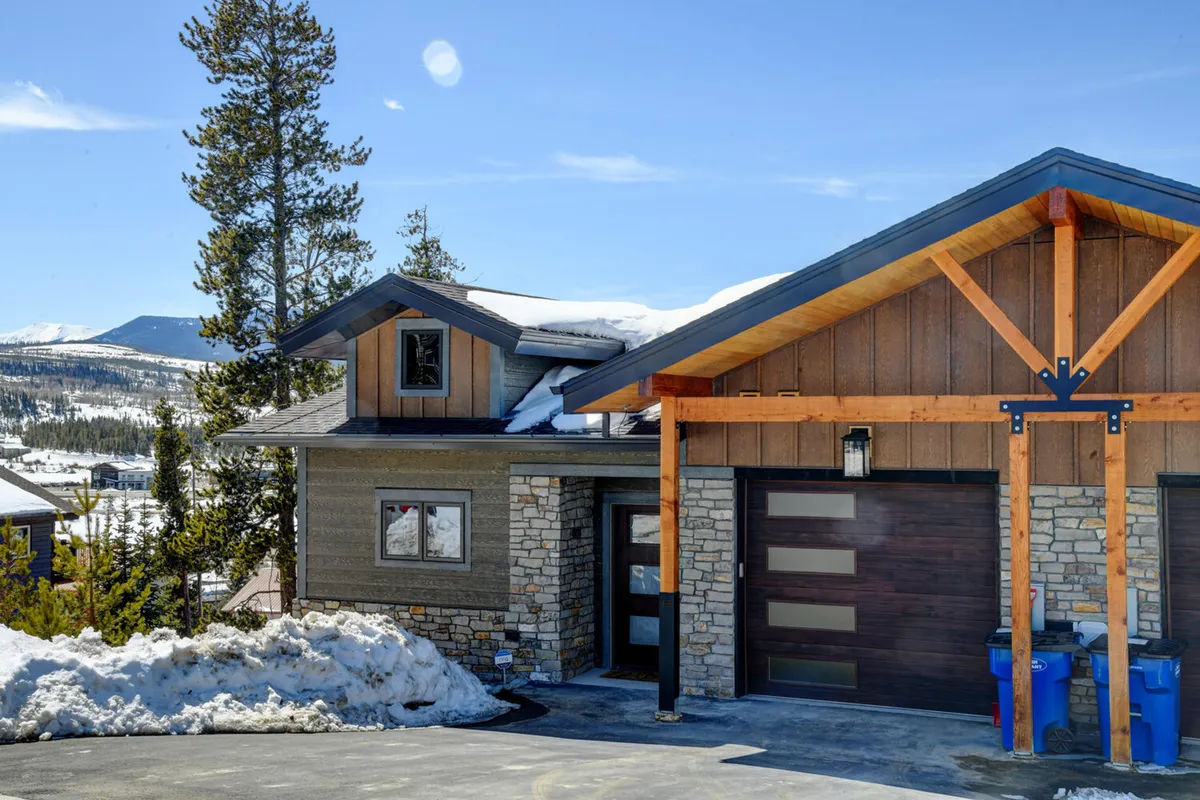 This image showcases the exterior architecture of the home, featuring a welcoming stone façade, wooden beams, and a modern garage door. Snow blankets the ground, and there's a sense of warmth from the porch, suggesting pet-friendly spaces and easy access to ski storage and garage parking.
