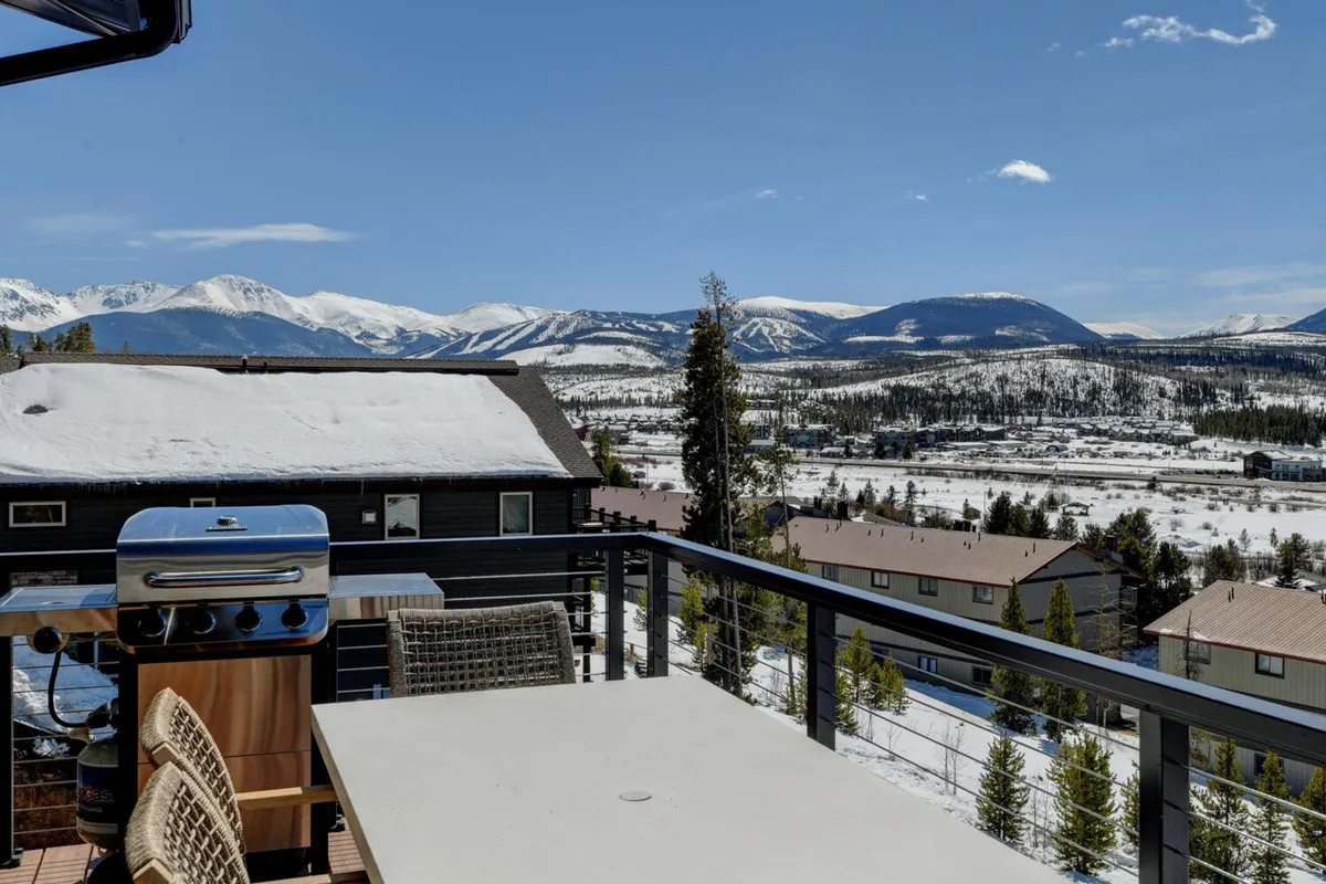 This photo presents an outdoor dining area set on a spacious deck, complete with a gas grill and a panoramic view of the mountain range. The setup shows a place for sunset dinners or snow-season barbecues, highlighting outdoor living amenities and year-round enjoyment.