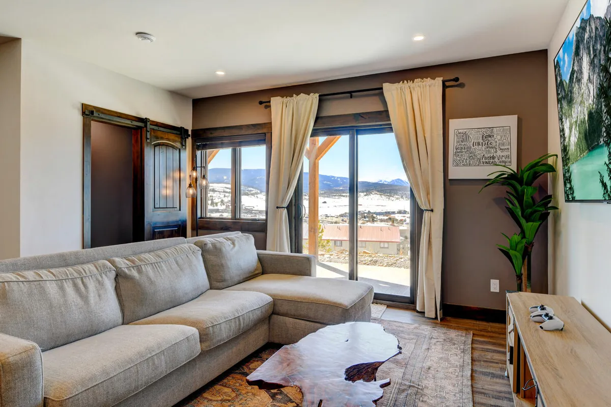 A spacious living area features a large sectional sofa, decorative area rug, an indoor plant, wall-mounted TV, and expansive windows looking out to the mountains. The space is ideal for group relaxation, game nights, or enjoying movies together, with nature just beyond the windows.