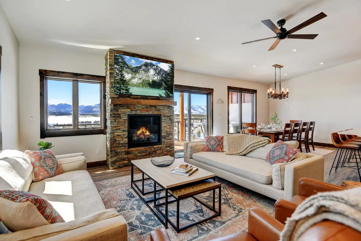 The image showcases a stunning interior living space featuring a crackling fireplace, expansive windows framing breathtaking mountain views, and a spacious dining area with comfortable seating—all set in a warm and welcoming atmosphere ideal for families or groups to gather, relax, and make memories.