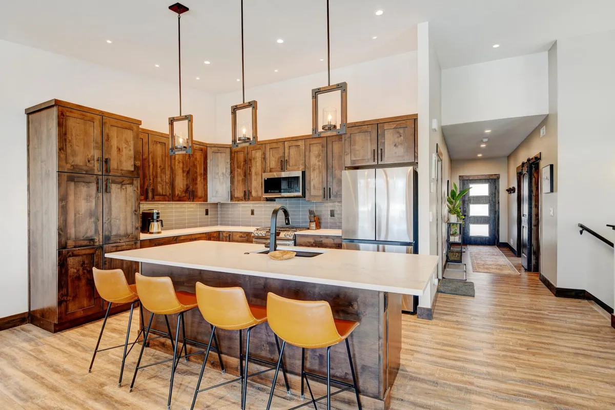 The contemporary chef’s kitchen is showcased with a prominent kitchen island complete with bar stools, sleek wooden cabinetry, modern stainless-steel appliances, and stylish lighting fixtures. The scene is inviting, highlighting a space designed for both culinary creativity and social interaction.